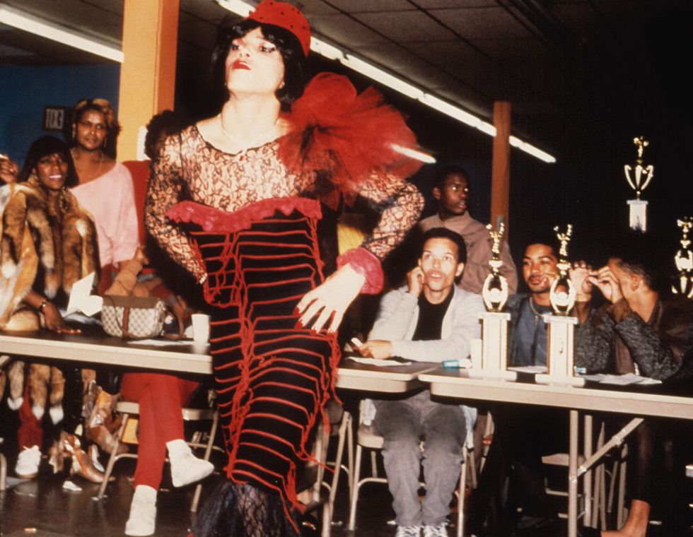 Particiant of a ball wearing a black dress with red accents walking on the stage, as the judges and the crowd watches them