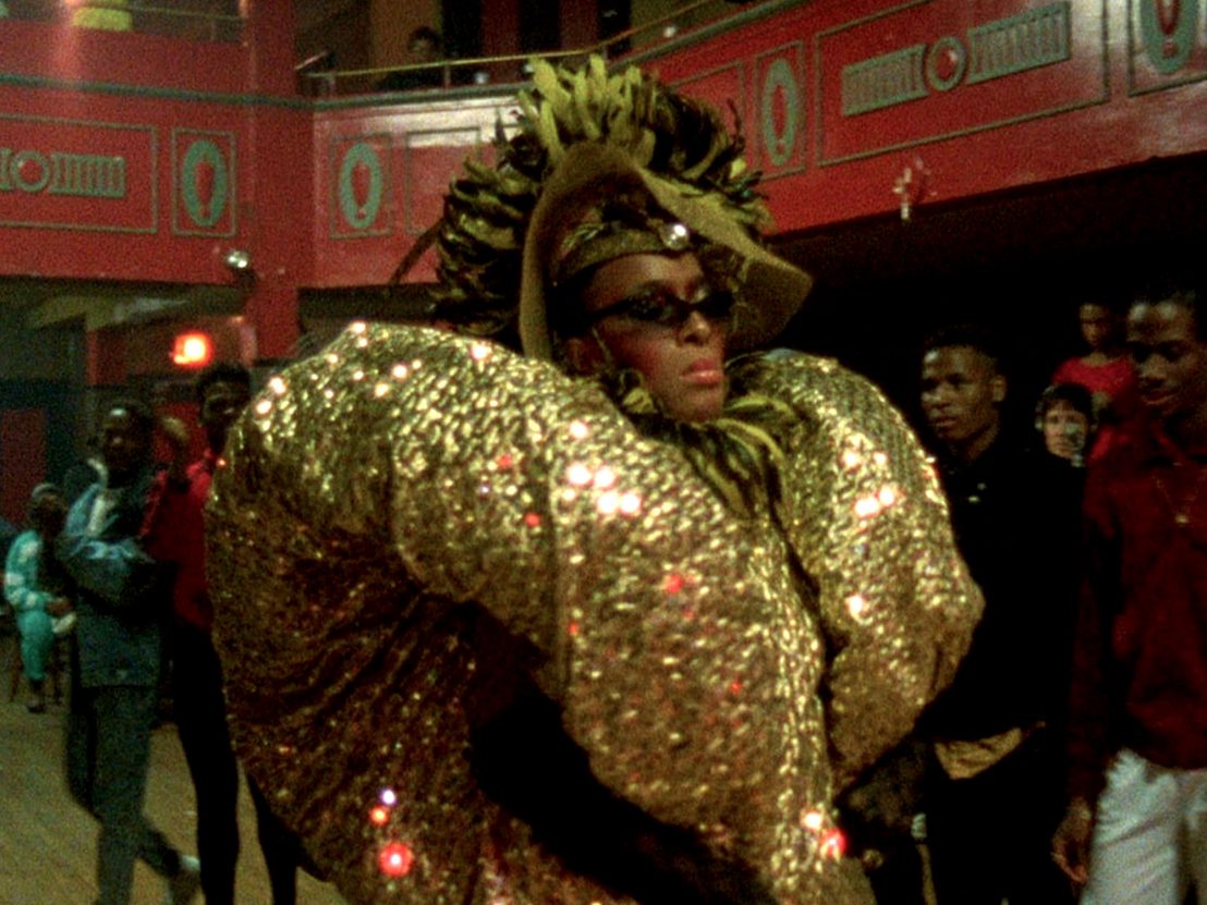 Black person wearing a gold shimering suit and headress, walking in a ballroom runway