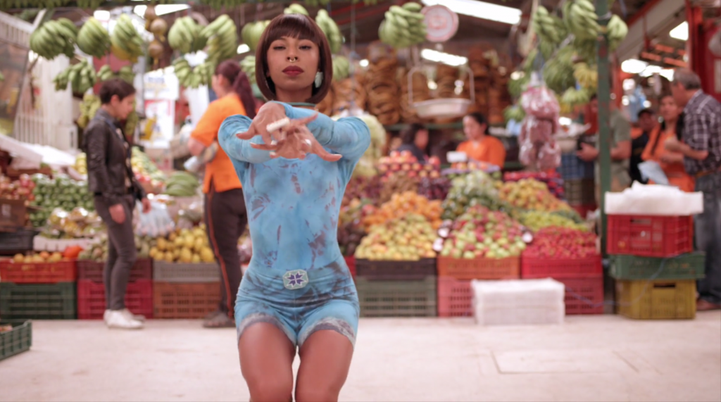 Kia LaBeija voguing in a public market, wearing a blue two pieces outfit, people and market shops in the background